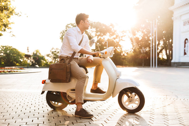Happy young businessman sitting on a motorbike outdoors, looking away - 写真・画像