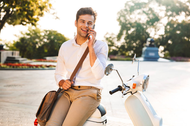 Happy young businessman sitting on a motorbike outdoors, talking on mobile phone - Photo, image