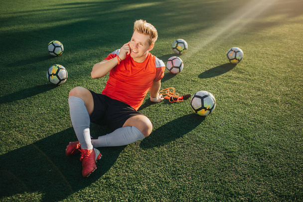 Relaxed young player lying on lawn and talk on phone. He looks straight. Guy keep legs crossed. Balls are behind him. - Foto, imagen