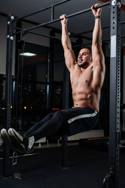 Brutal athletic man trains the muscles of the abdomen, hanging on the bar in the gym - Foto, immagini