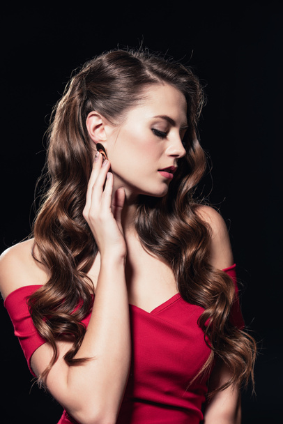 beautiful woman in red dress touching earring with hand and posing isolated on black - Foto, Imagem