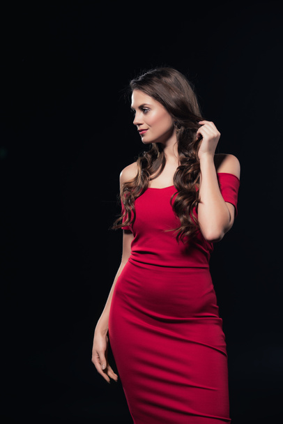 красивая женщина в красном платье касаясь волос, позируя и глядя в сторону изолированы на черном
 - Фото, изображение