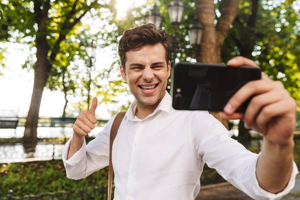 Happy young businessman wearing shirt taking a selfie while standing outdoors at the city park, holding mobile phone with outsretched hand - Foto, Bild