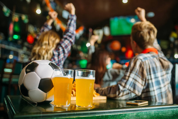 Ball and beer on the table in sports bar, football fans on background. Tv broadcasting, watching the game concept - Photo, Image