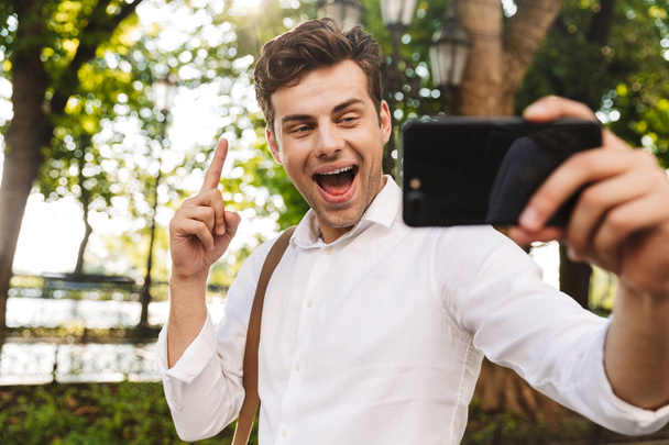 Happy young businessman wearing shirt taking a selfie while standing outdoors at the city park, holding mobile phone with outsretched hand - Photo, Image