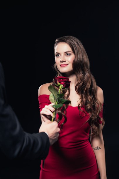 Mann präsentiert Rose an schöne lächelnde Frau in rotem Kleid isoliert auf schwarz - Foto, Bild
