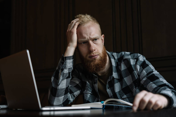 Young bearded hipster with emotional sad face feeling sick. Depression. Tired. Stress. Freelancer planning project, deadline. Frustrated businessman sitting in loft office Failure business, problems - Photo, Image