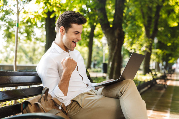 Cheerful young businessman sitting on a bench outdoors, working on laptop computer, celebrating success - Fotó, kép