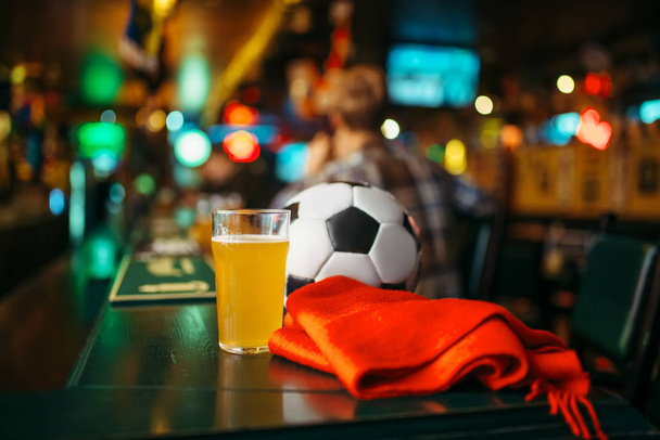 Ball, beer and red scarf on the counter in sports bar, football fans lifestyle. Tv broadcasting, watching the game concept - Photo, Image