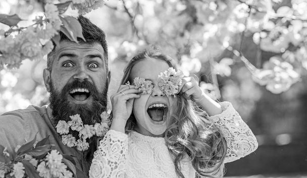 Father and daughter on happy face play with flowers as glasses, sakura background. Child and man with tender pink flowers in beard. Girl with dad near sakura flowers on spring day. Family time concep - Foto, Imagem