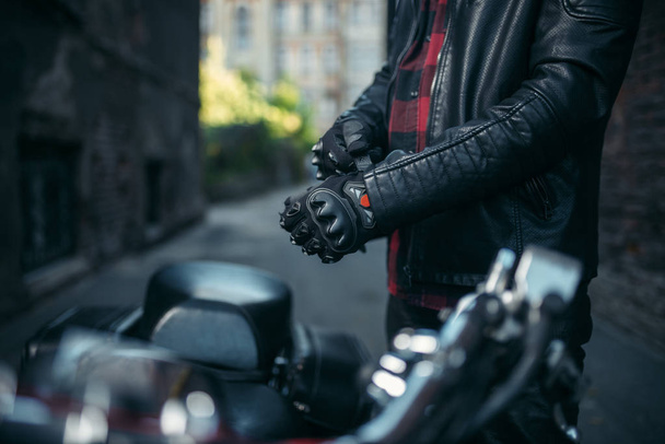 Male biker in leather jacket puts on gloves before riding on classical chopper, safety equipment - 写真・画像