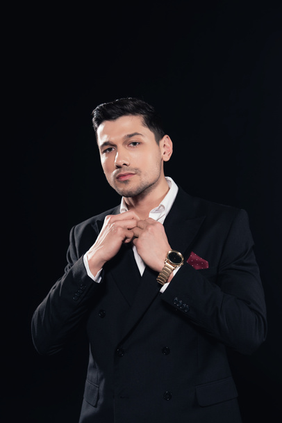 handsome man in suit looking at camera and buttoning up shirt isolated on black - Foto, imagen