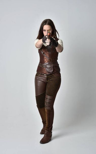 Ολόσωμο πορτρέτο του νέους, μελαχρινή κοπέλα φορώντας δερμάτινο ρούχο steampunk. πόζα στέκεται κρατώντας ένα όπλο σε φόντο γκρι στούντιο. - Φωτογραφία, εικόνα