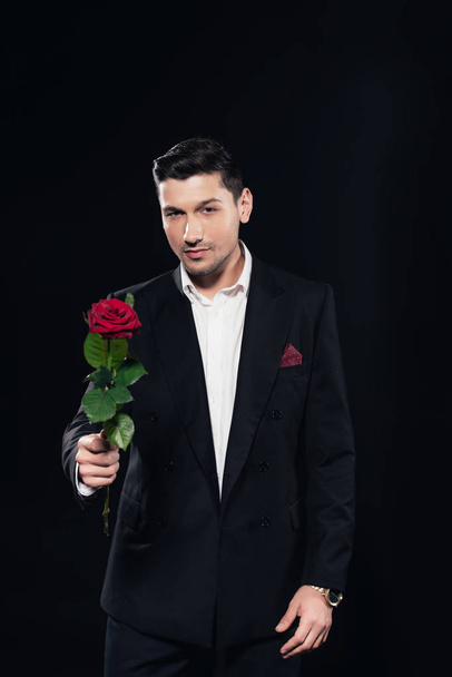 man in suit looking at camera and holding red rose isolated on black - Valokuva, kuva
