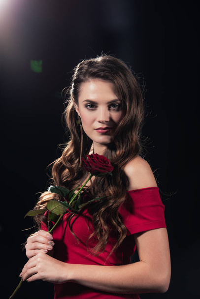 beautiful woman in red dress looking at camera and holding rose on black background - Foto, Bild