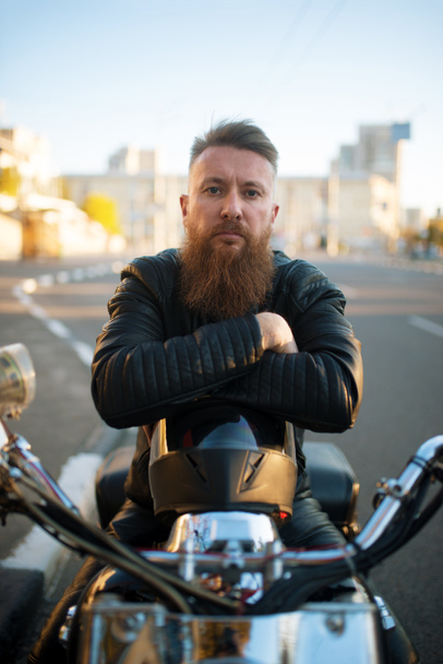 Bearded biker in leather jacket sitting on classical chopper, front view. Vintage bike, rider on motorcycle - Zdjęcie, obraz