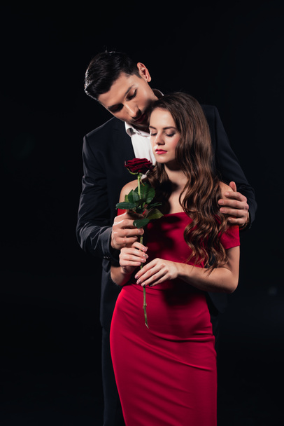 handsome man embracing beautiful woman in red dress holding rose isolated on black - Photo, Image