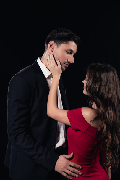 woman in red dress touching face of handsome man isolated on black - Foto, afbeelding
