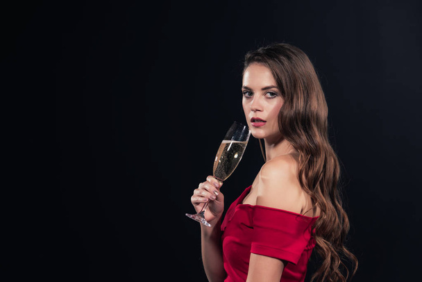 beautiful woman in red dress looking at camera and holding glass of champagne isolated on black - Foto, afbeelding