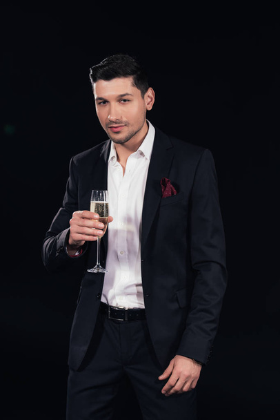 handsome man looking at camera and holding glass of champagne isolated on black - Foto, Imagem