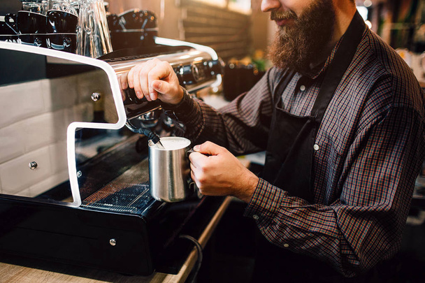 Young bearded barista floating milk. He does it careful. Guy wears apron. Young man prepares milk for coffee. - Foto, Bild