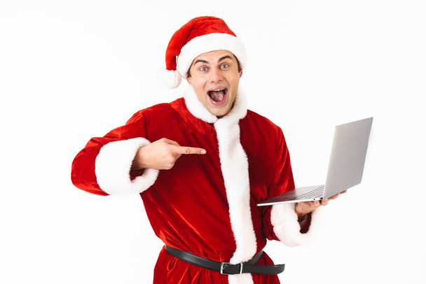 Portrait of a cheerful young man dressed in santa claus costume standing isolated over white background, holding laptop computer, pointing finger - Foto, immagini