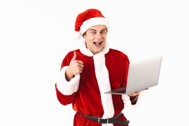 Retrato de um jovem alegre vestido em traje de Papai Noel de pé isolado sobre fundo branco, segurando computador portátil, mostrando polegares para cima
 - Foto, Imagem