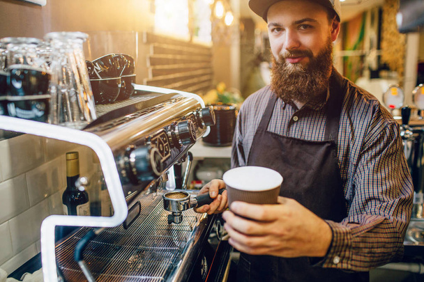 Young bearded man stand at coffee machine and hold cup of latte in hand. He looks on camera. - Fotoğraf, Görsel
