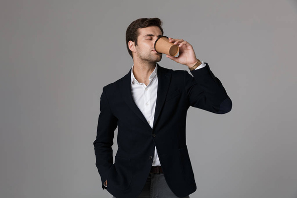 Imagen del hombre adulto de 30 años en traje de negocios bebiendo café de taza de papel aislado sobre fondo gris
 - Foto, Imagen