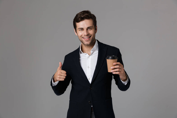 Image of attractive businessman 30s in suit smiling and holding paper cup with coffee isolated over gray background - Φωτογραφία, εικόνα