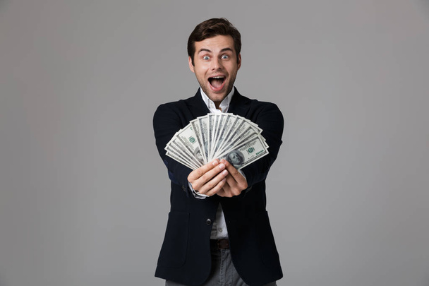 Image of joyful businessman 30s in suit smiling and holding fan of money in dollar banknotes isolated over gray background - Foto, Imagen