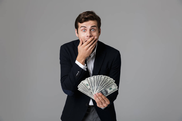 Image of surprised businessman 30s in suit smiling and holding fan of money in dollar banknotes isolated over gray background - Foto, afbeelding