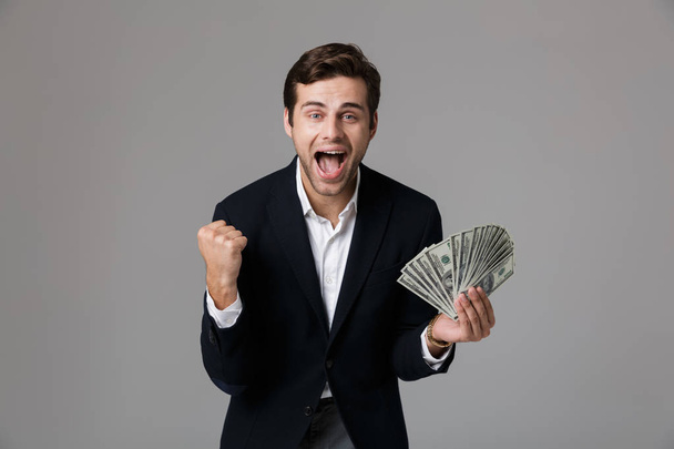 Image of joyous businessman 30s in suit smiling and holding fan of cash money isolated over gray background - Fotó, kép