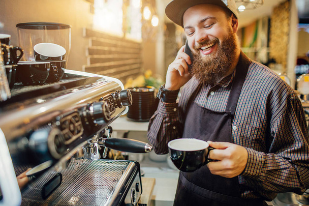 Nice bearded barista stands at coffee machine and talks on phone. He smiles. Guy hold cup of coffee in hands. - 写真・画像