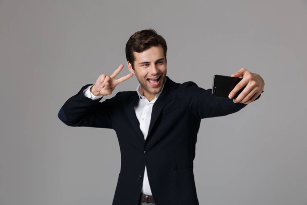 Obrázek optimistické podnikatel 30s ve formálním obleku, zobrazující znamení míru při pořizování selfie foto na mobil nad šedým pozadím, samostatný - Fotografie, Obrázek