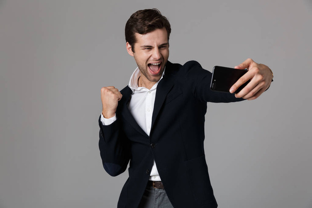 Image of cheerful man 30s in formal suit clenching fists while taking selfie photo on cell phone isolated over gray background - 写真・画像
