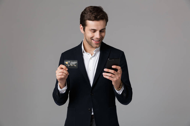 Image of successful businessman 30s in suit holding credit card and smartphone isolated over gray background - Valokuva, kuva
