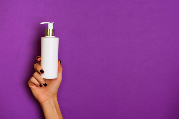 Female hands holding white cosmetic bottle on violet background. Banner. Skin care, pure beauty, body treatment concept. - Zdjęcie, obraz