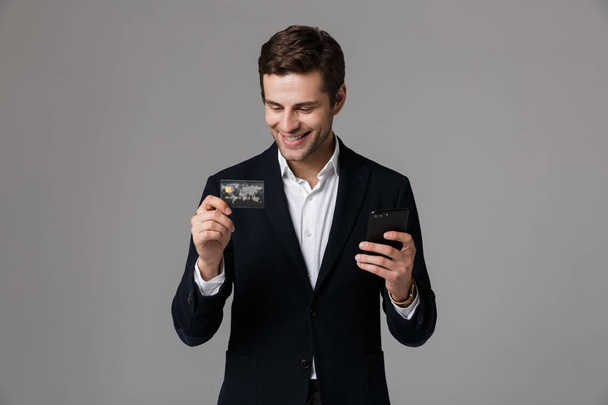 Image of cheerful businessman 30s in suit holding credit card and smartphone isolated over gray background - Фото, изображение
