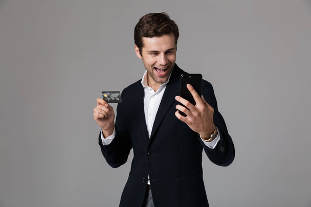 Portrét radostné mladý podnikatel oblečený v obleku izolované nad šedým pozadím, pomocí mobilního telefonu, ukazující kreditní kartu - Fotografie, Obrázek