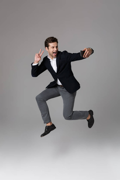 Full length portrait of a delighted young businessman dressed in suit isolated over gray background, taking a selfie while jumping - Foto, Bild