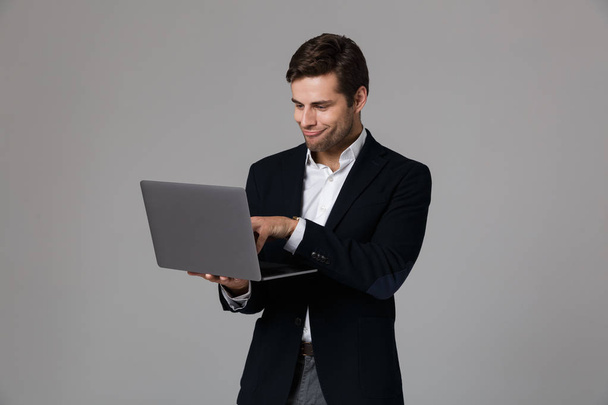 Image of happy businessman 30s in suit rejoicing while using laptop isolated over gray background - Φωτογραφία, εικόνα