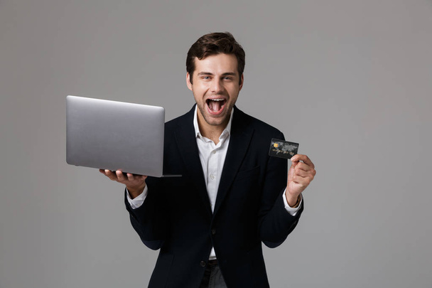 Image of delighted businessman 30s in suit screaming while holding laptop and credit card isolated over gray background - Fotoğraf, Görsel