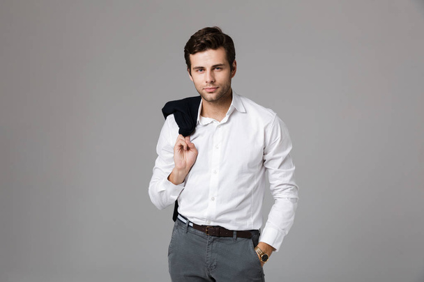 Close up portrait of a handsome young businessman standing isolated over gray background, looking at camera, holding a jacket - Фото, зображення