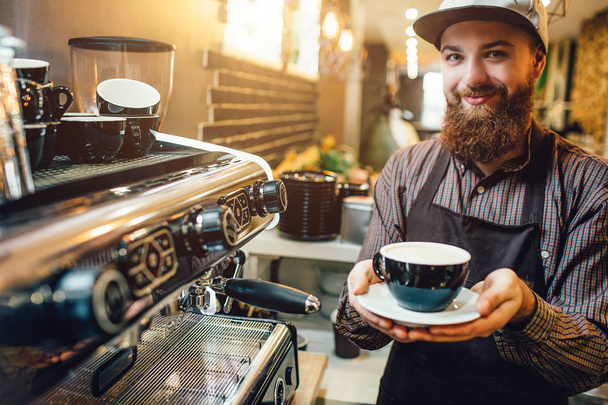 Veselé a příjemné barista držící šálek kávy v ruce. Ten stojí v kuchyni na kávovar. - Fotografie, Obrázek