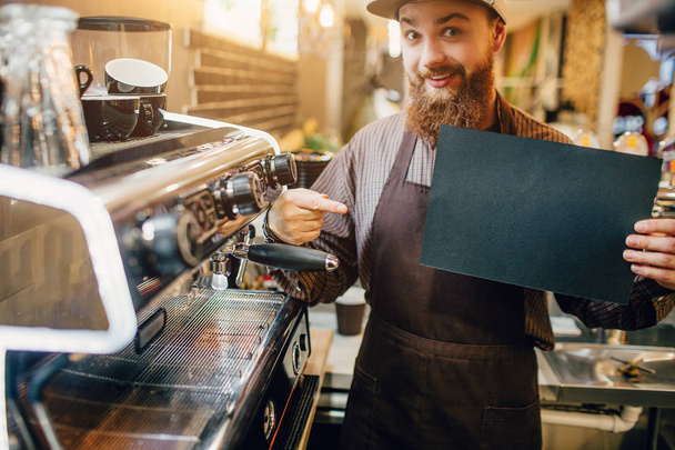 Excited young man points on piece of dark paper. He hold it in hand and look on camera. Guy stand at coffee machine in kitchen. - 写真・画像