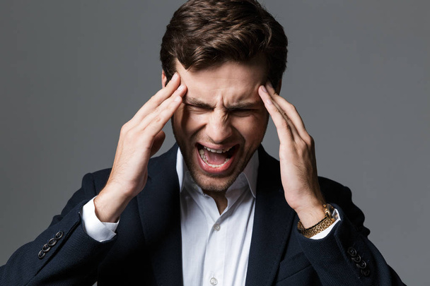 Close up portrait of a tired young businessman dressed in suit isolated over gray background, suffering from a headache, screaming - Photo, Image