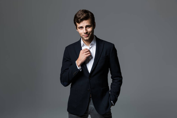 Portrait of a handsome young businessman dressed in suit isolated over gray background, unbuttoning his shirt - Foto, Imagem