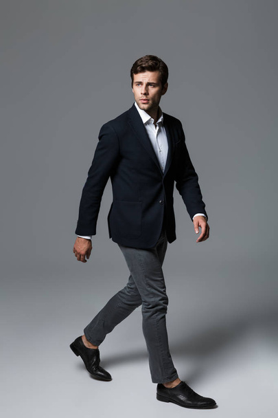 Full length portrait of a handsome young businessman dressed in suit isolated over gray background, posing, looking away, walking - Foto, Imagem