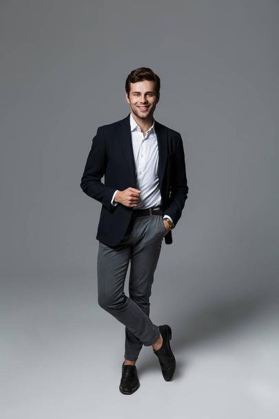 Full length portrait of a smiling young businessman dressed in suit isolated over gray background, posing, looking at camera - Фото, изображение